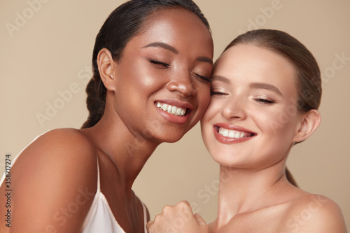 Beauty. Diversity Women Portrait. Happy Ethnic Models With Closed Eyes Standing Together Against Beige Background. Tender Caucasian And Mixed Race Female With Perfect Skin And Natural Makeup.