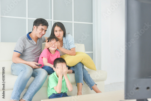 Asian family sitting on sofa and watching TV together.Mother closing daughter eyes by hands and son closing eyes by himself. Violence on TV programe concept.