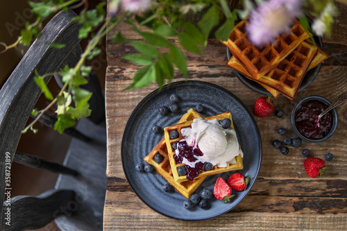 Homemade waffles with ice cream, fresh berries and jam on blue plate. Rustic wooden table. Traditional Belgian waffles. Close up view, copy space.  Beautiful dessert or breakfast, country style.
