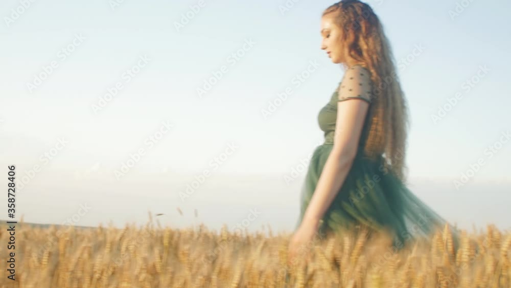 Beautiful woman in green dress walkink across field and touches ears of wheat with hand at sunset light, girl enjoying summer nature landscape Beautiful woman in green dress walkink across field and touches ears of wheat with hand at sunset light, girl enjoying summer nature landscape