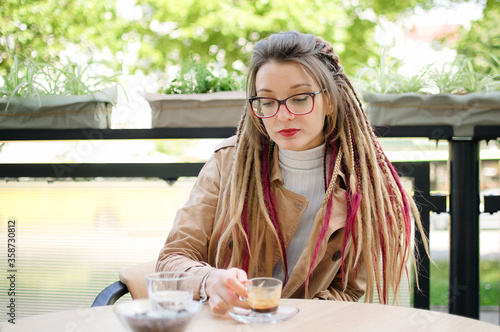 Young woman wearing glasses, casual nude trench and long dreadlocks is drinking coffee in outdoor cafe.