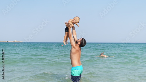 Young dad standing in water in sea and throw up smiling little son in her arms