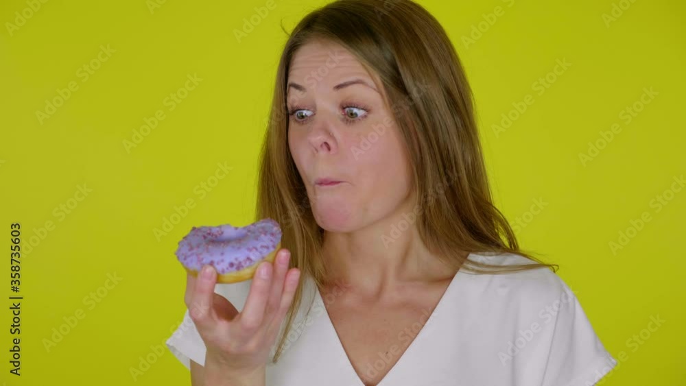 Woman in a white T-shirt bites, eats a delicious blue donuts and enjoys the taste on a yellow background with copy space. Place for text or product. 4K slow motion footage Woman in a white T-shirt bites, eats a delicious blue donuts and enjoys the taste on a yellow background with copy space. Place for text or product. 4K slow motion footage