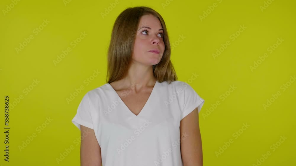 Woman in a white T-shirt looks around and thinks what to choose on a yellow background with copy space. Place for text or product. 4K slow motion footage Woman in a white T-shirt looks around and thinks what to choose on a yellow background with copy space. Place for text or product. 4K slow motion footage