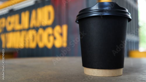 black paper cup without text and logo with coffee with a plastic cover on a wooden table of a street coffee shop in the city. Copy space.