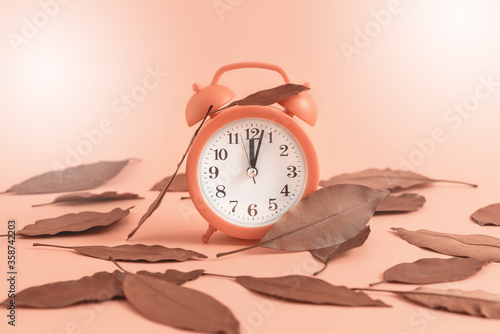 Alarm clock among autumn fallen leaves on pastel faded background.