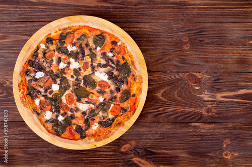 Delicious pizza with cherry tomatoes, spinach, mozzarella, feta, kalamata olive and mushrooms on a round wood platter which is on the wooden rustic table, top view with copy space