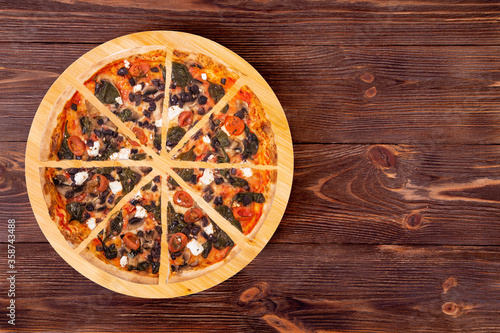 Sliced pizza with cherry tomatoes, spinach, mozzarella, feta, kalamata olive and mushrooms on round wood plate which is on wooden rustic table, top view and space for your text