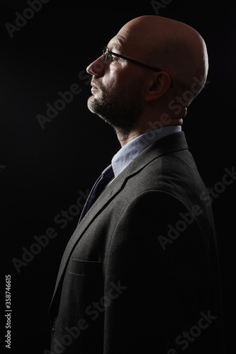 Dark dramatic portrait of a bald slim businessman in grey suit, blue shirt, tie and glasses. Profile view of the face. Black background.