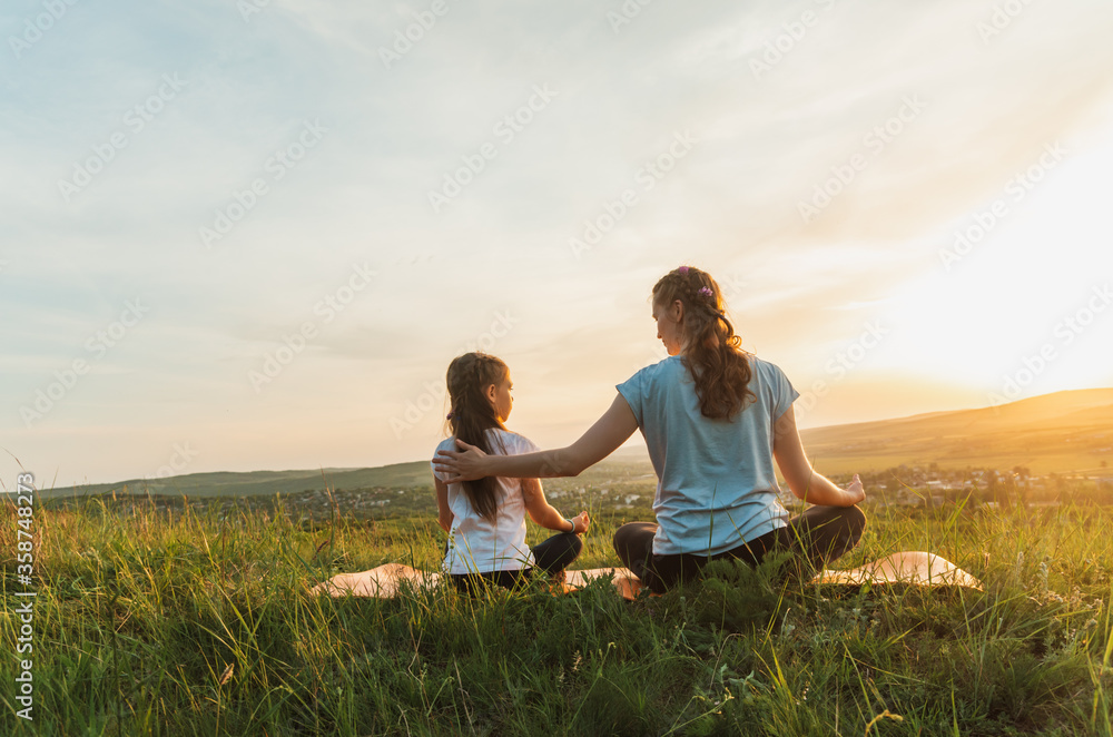 Back view of young woman and child sitting cross-legged on a hill overlooking rural landscape, at sunset 