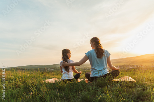 Back view of young woman and child sitting cross-legged on a hill overlooking rural landscape, at sunset 