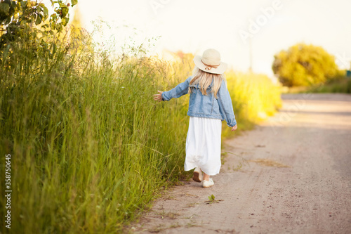 A cute girl in a denim jacket and a white dress in a straw hat is walking along the road. Back view. The concept of summer, happy childhood, freedom.