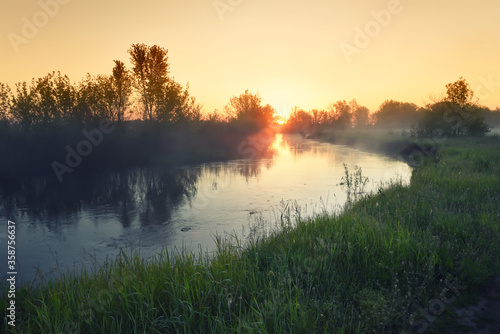 Dawn on the lake. The sun in the fog above the water and grass on the shore. 