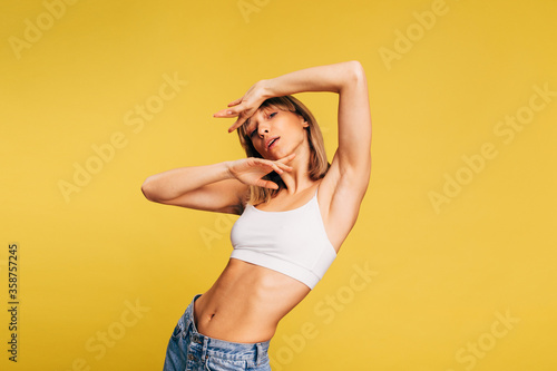 Gorgeous flexible young woman posing on camera with full body. Shows her amazing abs muscles on naked stomach. Model in white crop top and jeans isolated over yellow background.