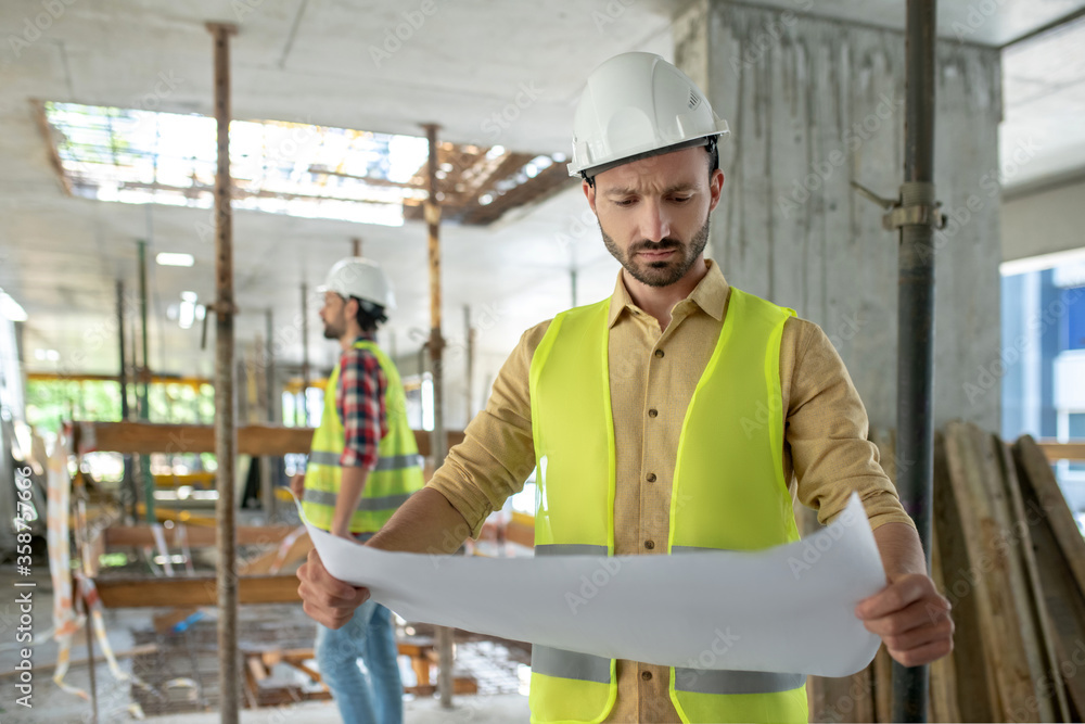 Building worker in yellow vest studying blueprint, his colleague standing in the background, checking construction Building worker in yellow vest studying blueprint, his colleague standing in the background, checking construction