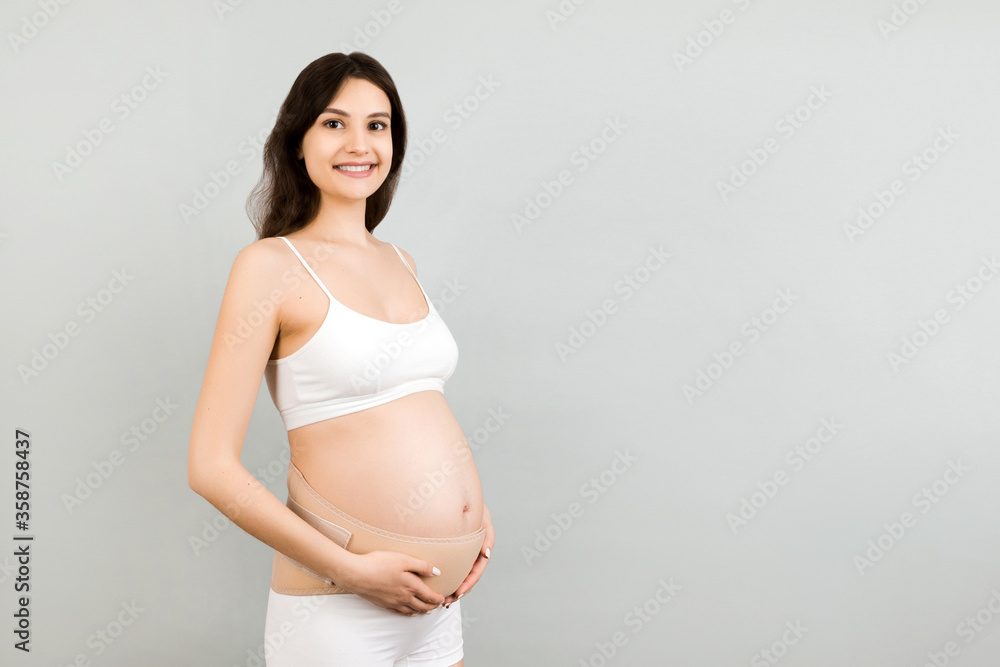 Portrait of pregnant woman in underwear wearing maternity belt to reduce  pain in the back at gray background with copy space. Orthopedic abdominal  support belt concept Stock Photo