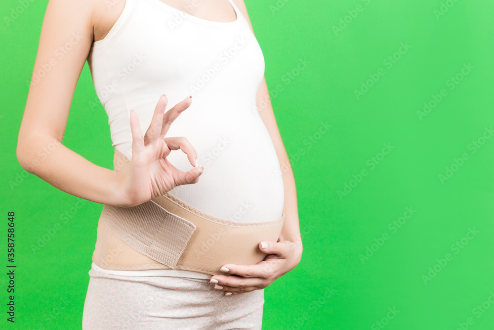Close up of pregnant woman wearing supporting bandage and showing okay gesture at green background with copy space. Orthopedic abdominal support belt concept Close up of pregnant woman wearing supporting bandage and showing okay gesture at green background with copy space. Orthopedic abdominal support belt concept