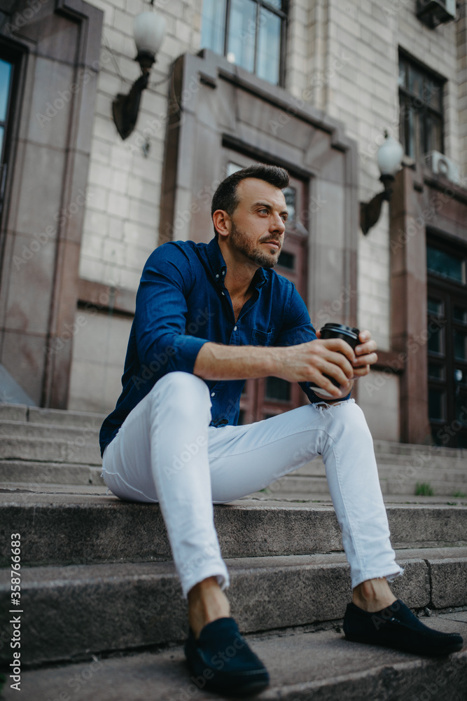 Young man sits and ponders on stairs with cup of coffee near building. Young man sits and ponders on stairs with cup of coffee near building.