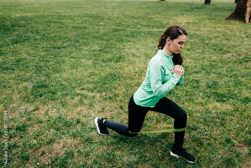 Side view of attractive sportswoman doing lunges with resistance band in park