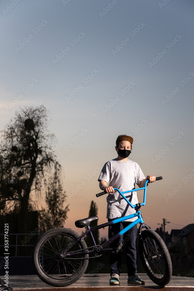 Fototapeta premium Portrait of a teenager in the sunset in a black mask on a blue bicycle. Quarantine. It is forbidden to ride a bicycle. Covid-19.