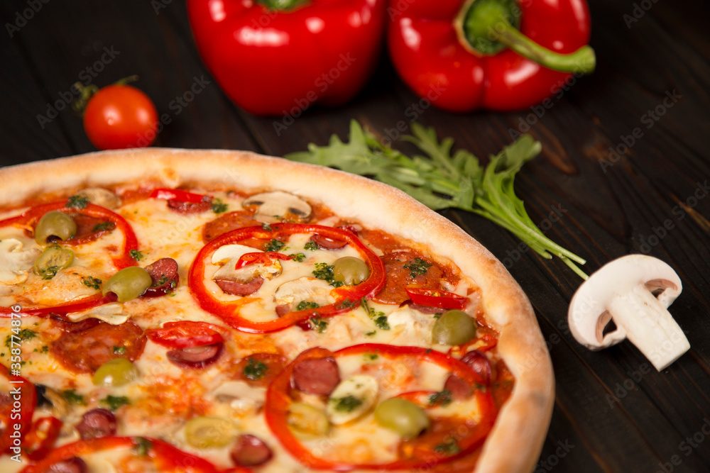 Side view of pizza with bell pepper, green olives, smoked sausages, mozzarella cheese, pepperoni, salami, chili pepper, champignons on wooden board with two bell peppers and herbs on background
