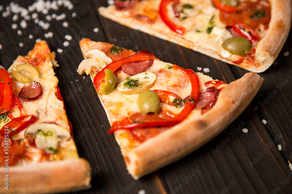 Slice of delicious pizza. Side view of pizza with bell pepper, olives, sausages, mozzarella cheese, pepperoni, salami, chili pepper, champignons on wooden background with sea salt. Close up of piece
