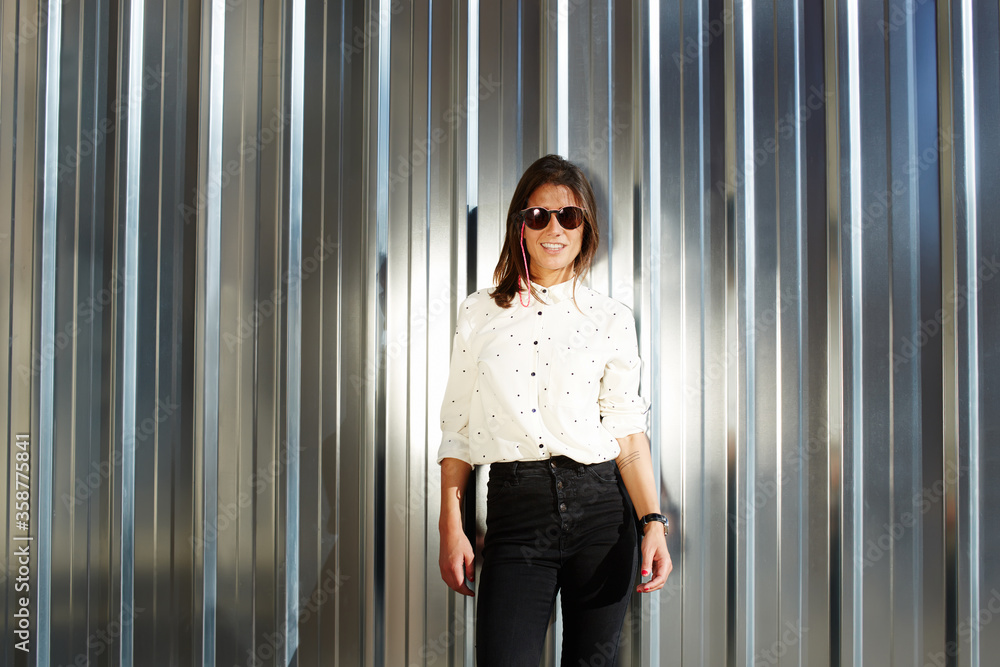 Glamorous female model in black sunglasses posing for the camera outdoors while standing on chrome shiny wall background with copy space area, fashionable young hipster girl with smile looking to you Glamorous female model in black sunglasses posing for the camera outdoors while standing on chrome shiny wall background with copy space area, fashionable young hipster girl with smile looking to you