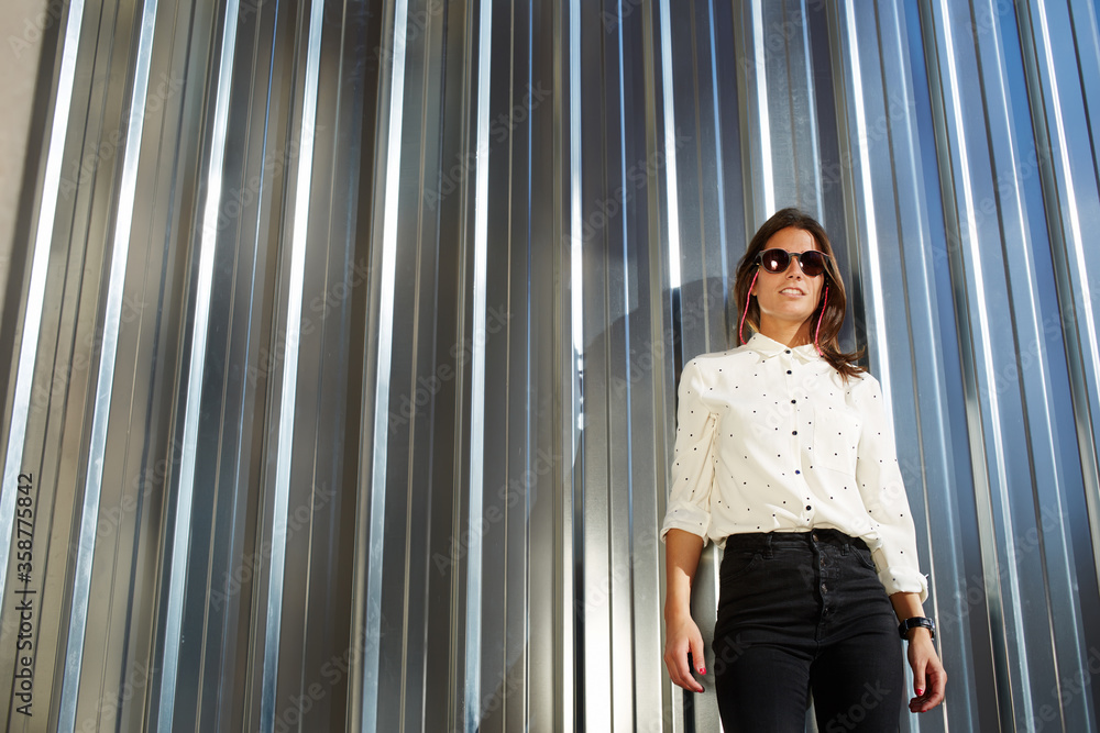 Young fashionable woman in trendy clothing and black sunglasses standing on metallic wall background with copy space for your text message or advertising content, female hipster dressed with style Young fashionable woman in trendy clothing and black sunglasses standing on metallic wall background with copy space for your text message or advertising content, female hipster dressed with style