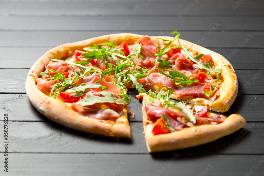 Close up of traditional Italian pizza with jamon, arugula and cherry tomatoes on dark wooden background. Slice of pizza moved near the whole dish. Delicious Mediterranean meal with fresh herbs
