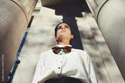 Low view portrair of a young beautiful woman with stylish clothes and classy modern sunglasses standing near a large building with conceived serious face looking afield, female posing for the camera photo