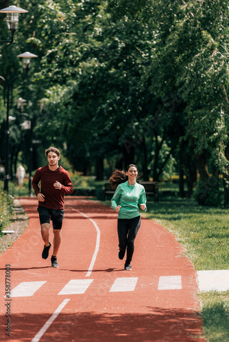 Fototapeta Naklejka Na Ścianę i Meble -  Smiling woman jogging near handsome boyfriend on running track in park