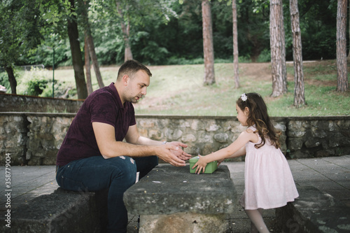 Side view of father and daughter in park. Little daughter sitting on stone bench with her father and giving him a present.