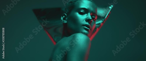 Cute girl model illuminated by colorful neon light poses in studio, emotionally looking at camera. Portrait of beautiful young woman with shiny metallic makeup on face and body. Fashion model shooting