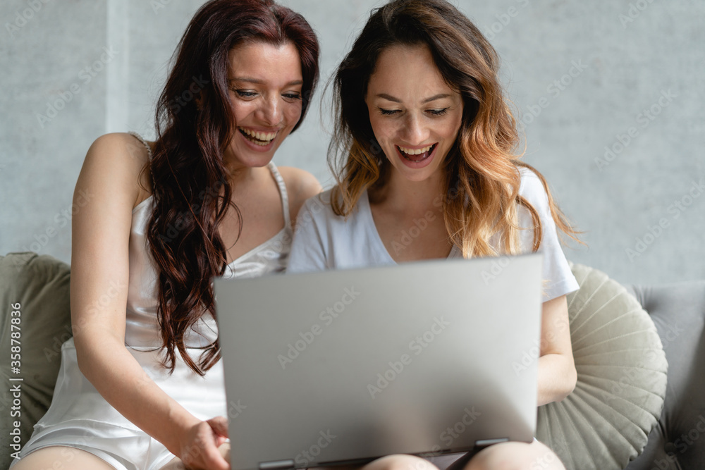Two young Caucasian beautiful sisters are sitting in a room on the couch and are looking at the laptop screen. They are smiling brunettes with loose hair and white pajamas. Two young Caucasian beautiful sisters are sitting in a room on the couch and are looking at the laptop screen. They are smiling brunettes with loose hair and white pajamas.