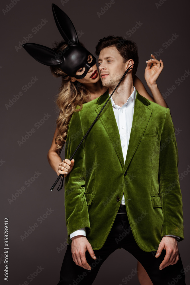 Woman in rabbit mask holding flogging whip near handsome boyfriend isolated on grey
