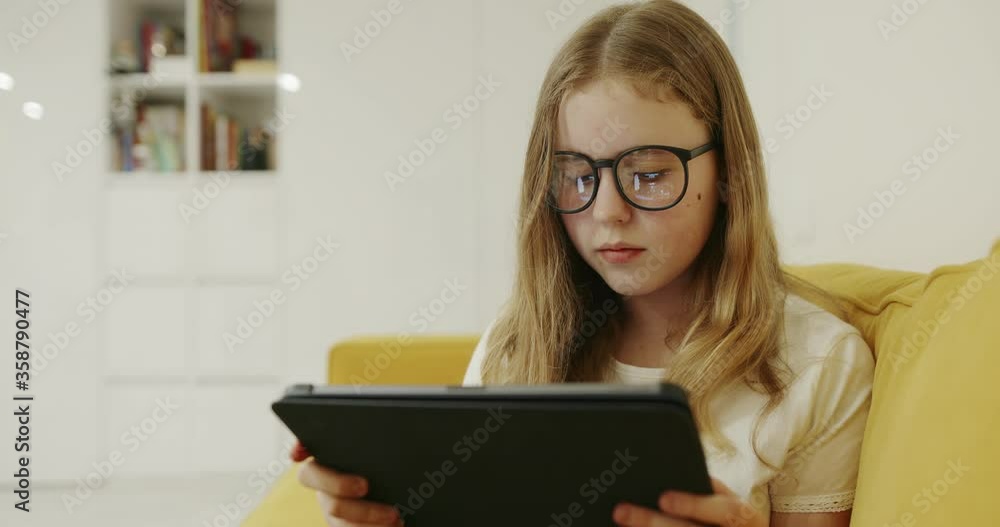 Smart girl in glasses sitting on couch and playing game on tablet while spending time at home during quarantine Smart girl in glasses sitting on couch and playing game on tablet while spending time at home during quarantine
