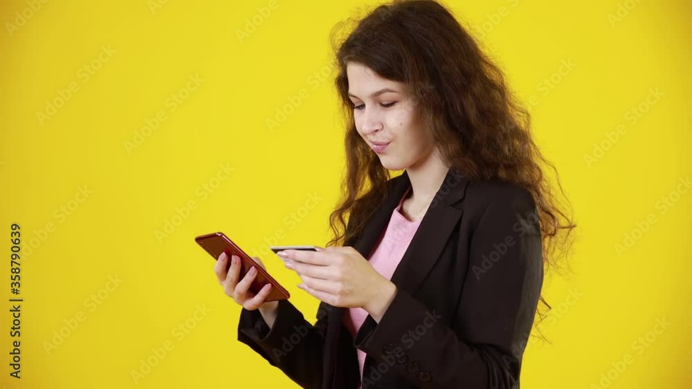 Satisfied caucasian pretty lady using credit card and making online order, paying for purchases with her smartphone on yellow background wall. Concept of modern technology. Satisfied caucasian pretty lady using credit card and making online order, paying for purchases with her smartphone on yellow background wall. Concept of modern technology.