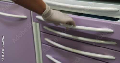 Hand with medical gloves opens drawer with clean white paper tissues, medical masks, and saliva ejectors inside, picks up one saliva suction tube and closes the drawer. Dentistry tools.