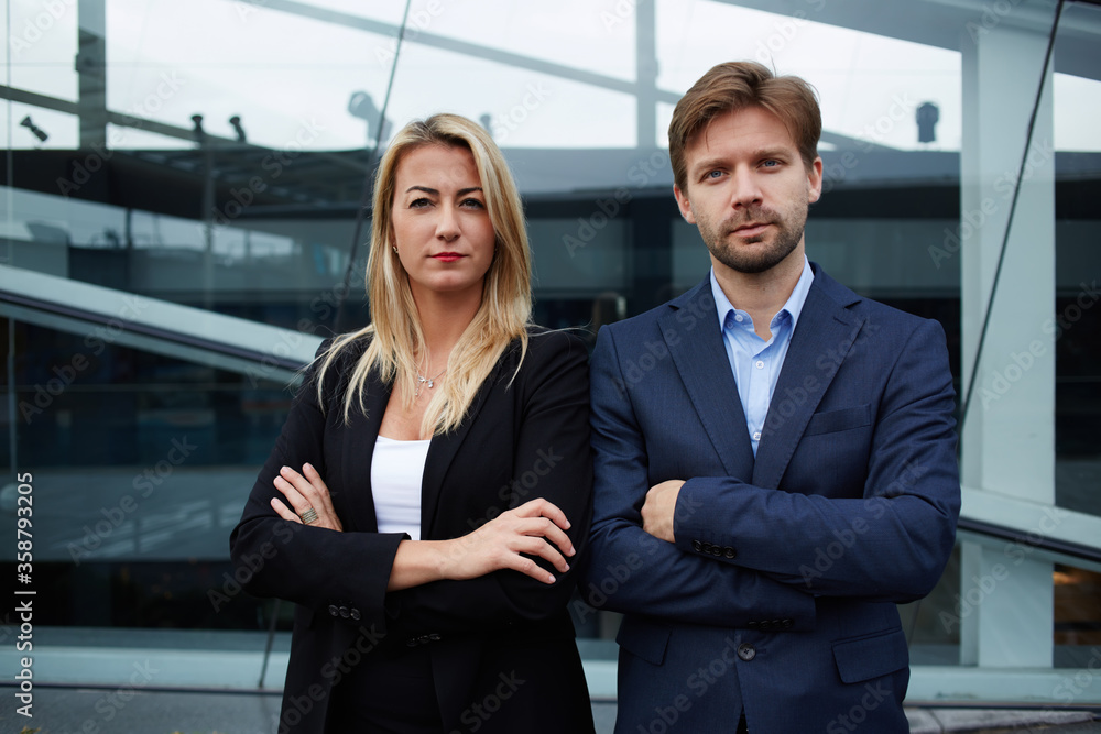 Two successful businesspeople crossing their arms while standing near office building, couple of confident work colleagues standing with their arms folded back to back, teamwork companions concept Two successful businesspeople crossing their arms while standing near office building, couple of confident work colleagues standing with their arms folded back to back, teamwork companions concept