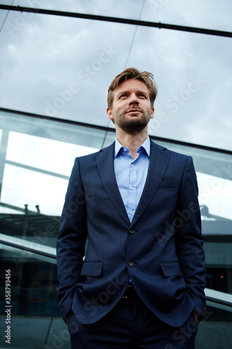 Low angle portrait of a handsome smiling businessman standing with his hands in his pockets in front of office building, carefree future vision concept
