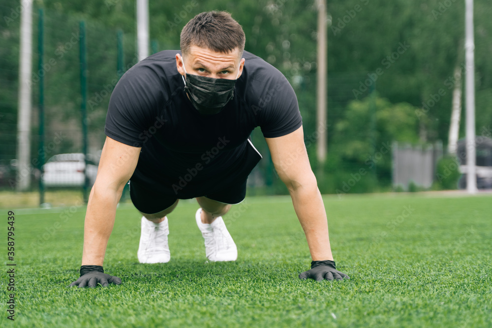 A young man in medical mask and disposable gloves makes push-ups on the grass in the fresh air. A young man in medical mask and disposable gloves makes push-ups on the grass in the fresh air.