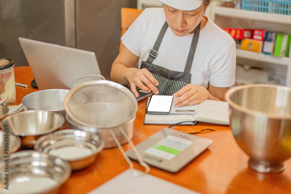 Female baker using cell phone with laptop and bakery ingredient on table. Female baker using cell phone with laptop and bakery ingredient on table.