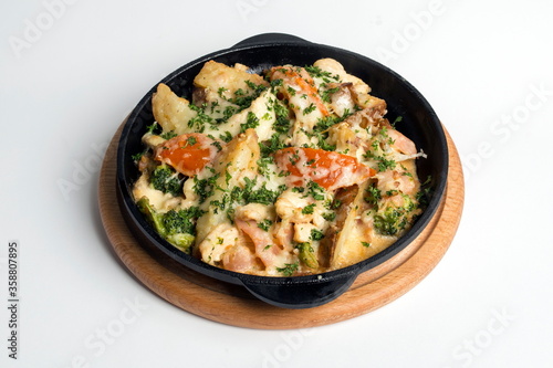 Dish on a black pan, which stands on a wooden stand. Potatoes with cheese and red pepper. Sprinkled with green parsley.