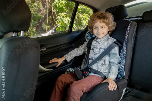 Happy cute boy of elementary age in denim jacket and brown pants sitting in car