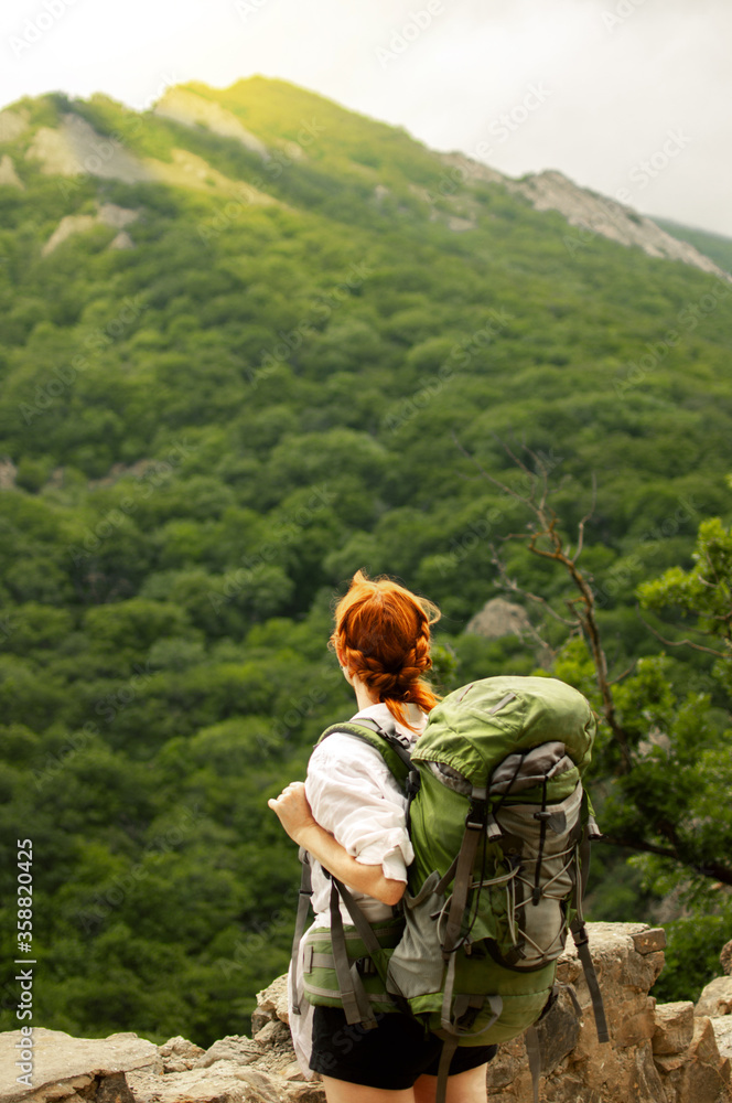A girl with a green backpack stands and looks at the mountains and forest. Travel concept, epic beautiful moment.