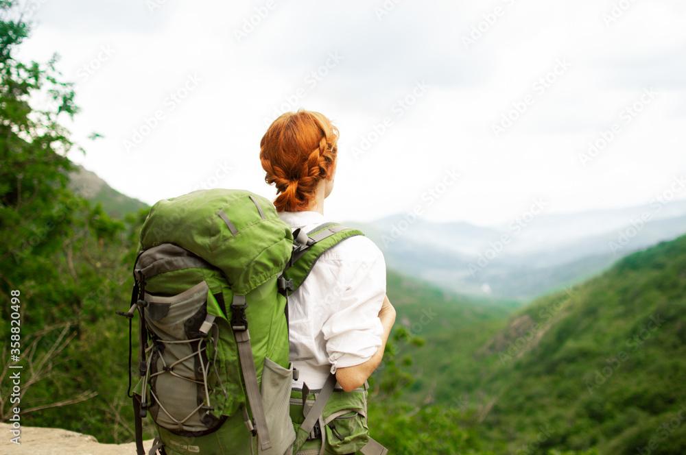 A girl with a green backpack stands and looks at the mountains and forest. Travel concept, epic beautiful moment.