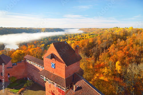 Close-up of the Turaida castle tower in a clouds of morning fog at sunrise. Colorful red, orange and yellow trees of the autumn forest. Gauja national park, Latvia. Travel guide, sightseeing theme photo