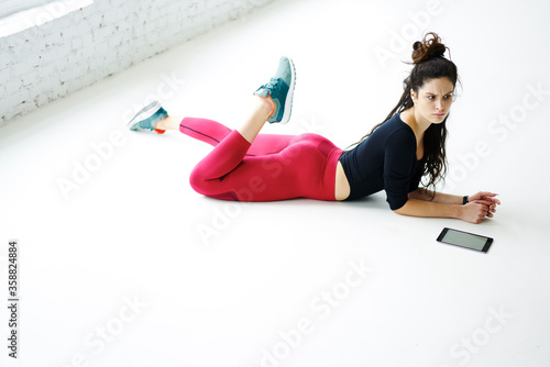 Attractive young female fitness instructor dressed in stylish sportswear recreating after workout in gym lying on white floor copy space for advertising using application touchpad counting activities