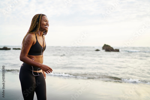 Fototapeta Naklejka Na Ścianę i Meble -  Cheerful african female runner dressed in sport top listening favourite music songs in modern earphones during sport workout walking on sea coastline of tropical island, concept of healthy lifestyle