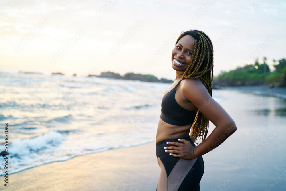 Portrait of cheerful afro american jogger with dreads and sport clothes posing at camera during morning training on seashore.Positive dark skinned female dressed in active wear standing on coastline Portrait of cheerful afro american jogger with dreads and sport clothes posing at camera during morning training on seashore.Positive dark skinned female dressed in active wear standing on coastline